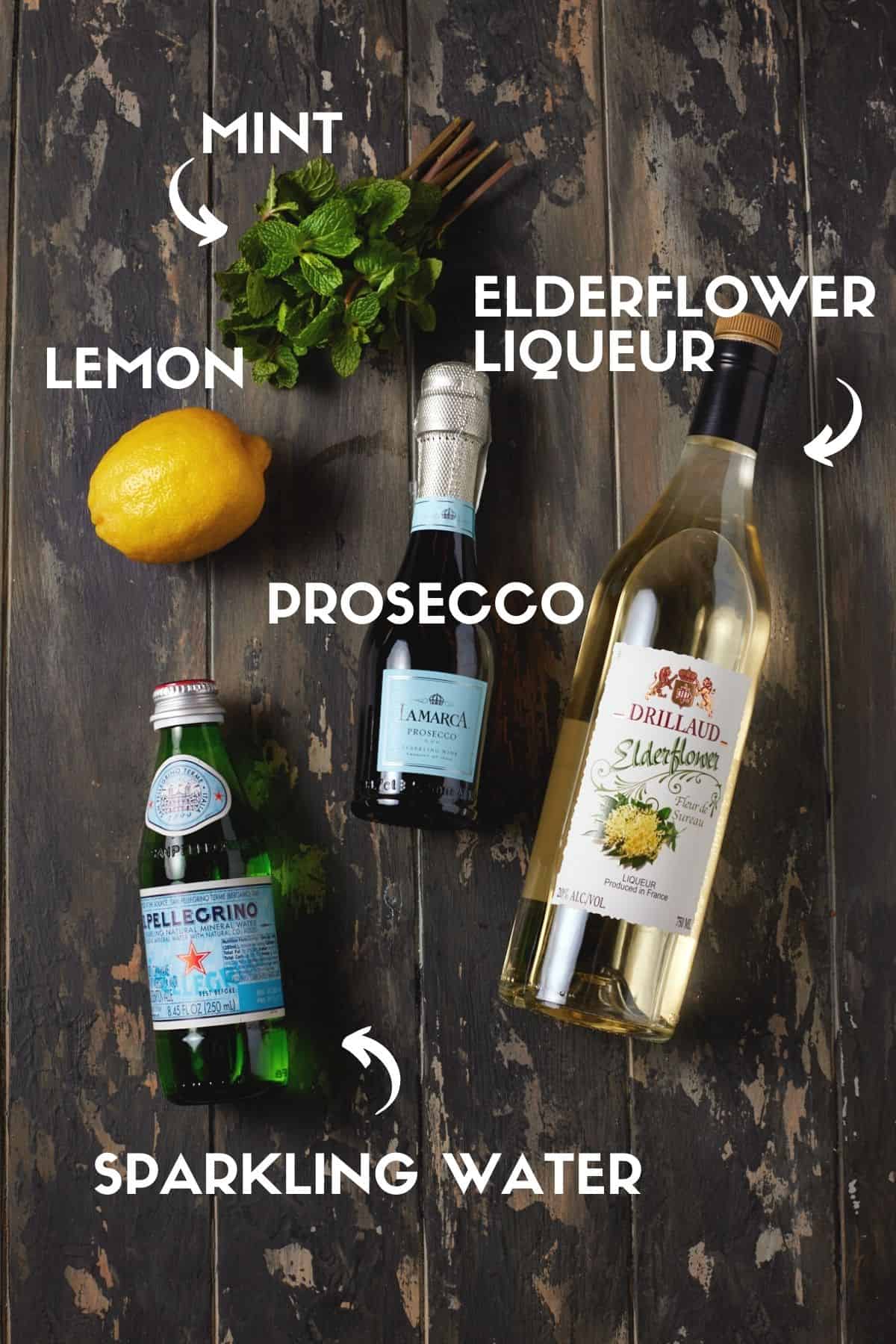 Bottles of prosecco, sparkling water, elderflower liqueur, a lemon and mint leaves on a wood board. 