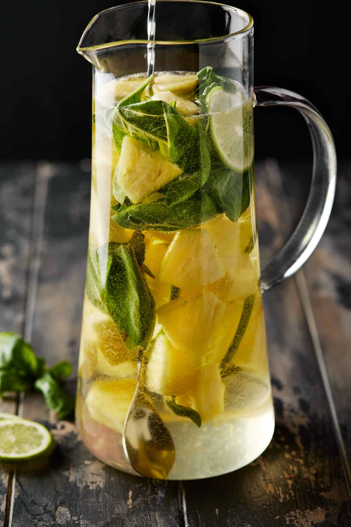 Glass pitcher of white wine sangria with pineapple, basil leaves and lime slices. 
