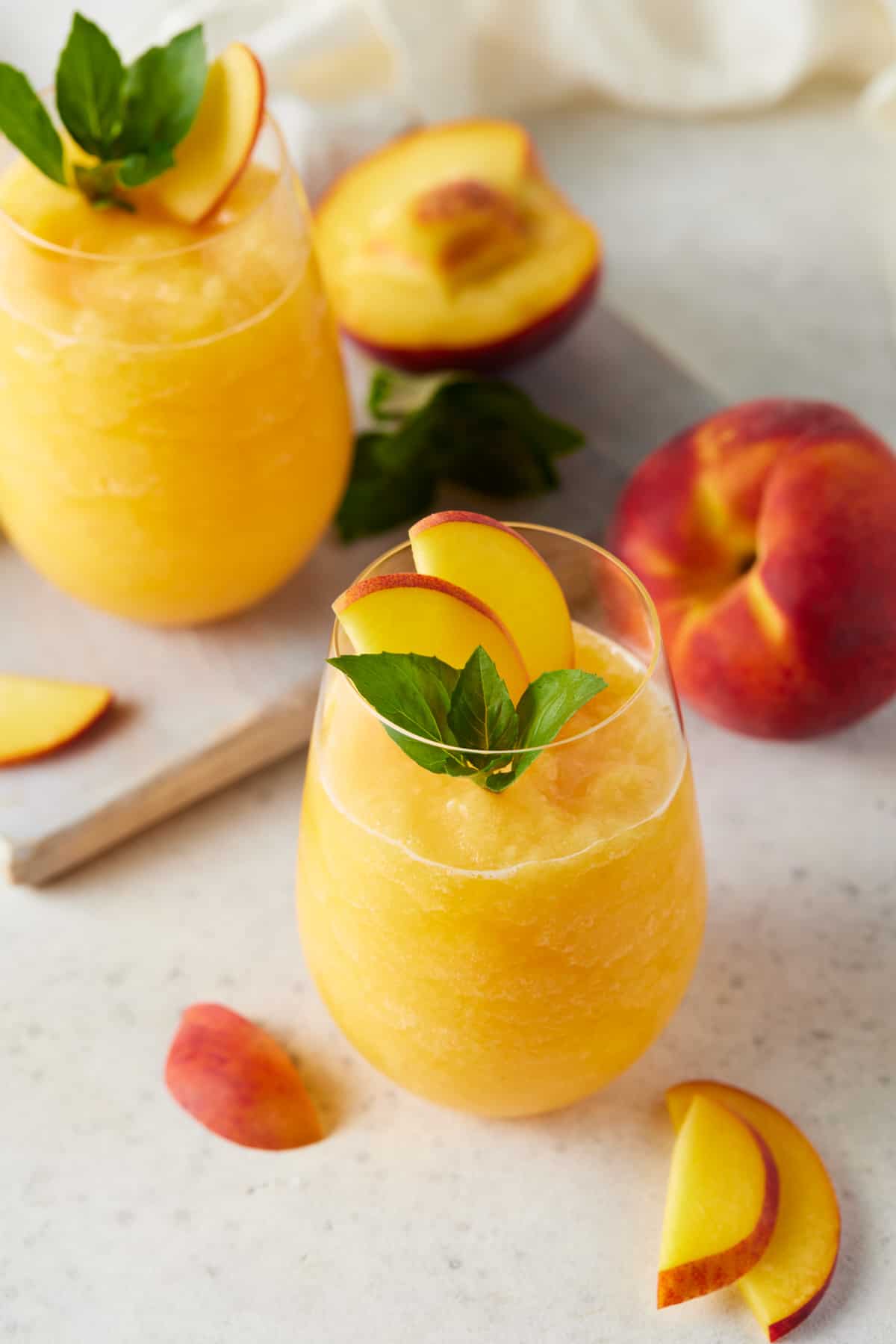 Two stemless wine glasses of peach wine slushies with fresh peach slices and fresh basil. 