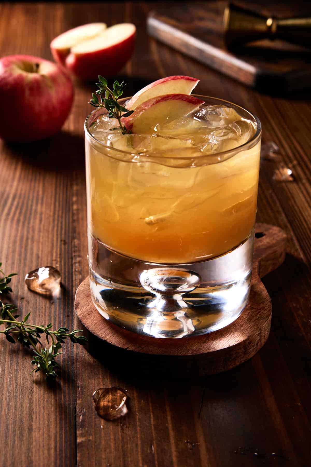 An apple cider bourbon cocktail on a dark wood board garnished with apple slices and fresh thyme. 
