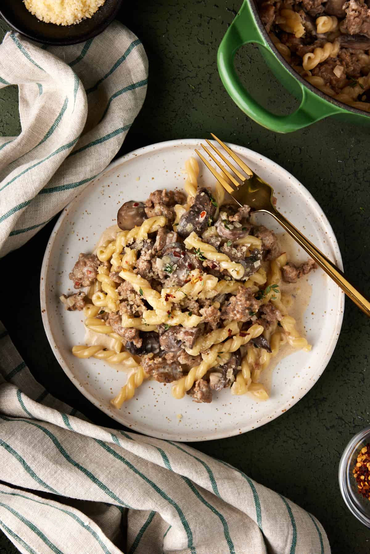 Cream plate of creamy sausage gemelli pasta with a green and beige striped towel. 