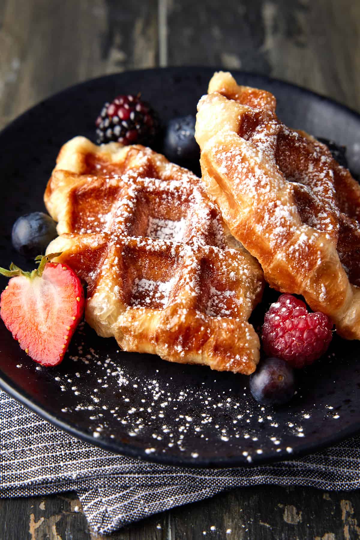 Two croffles with powdered sugar and berries on a black plate. 