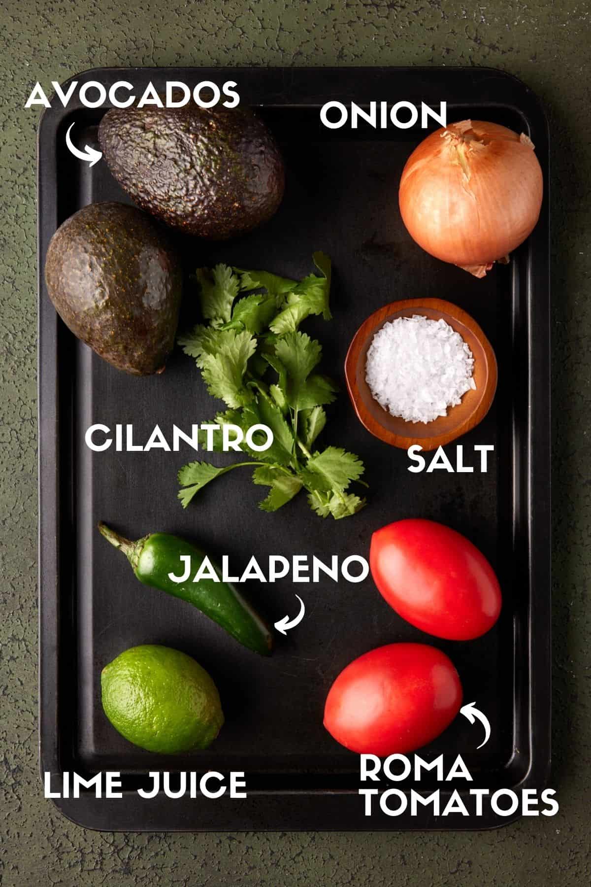 Guacamole ingredients on black sheet pan including avocados and tomatoes. 