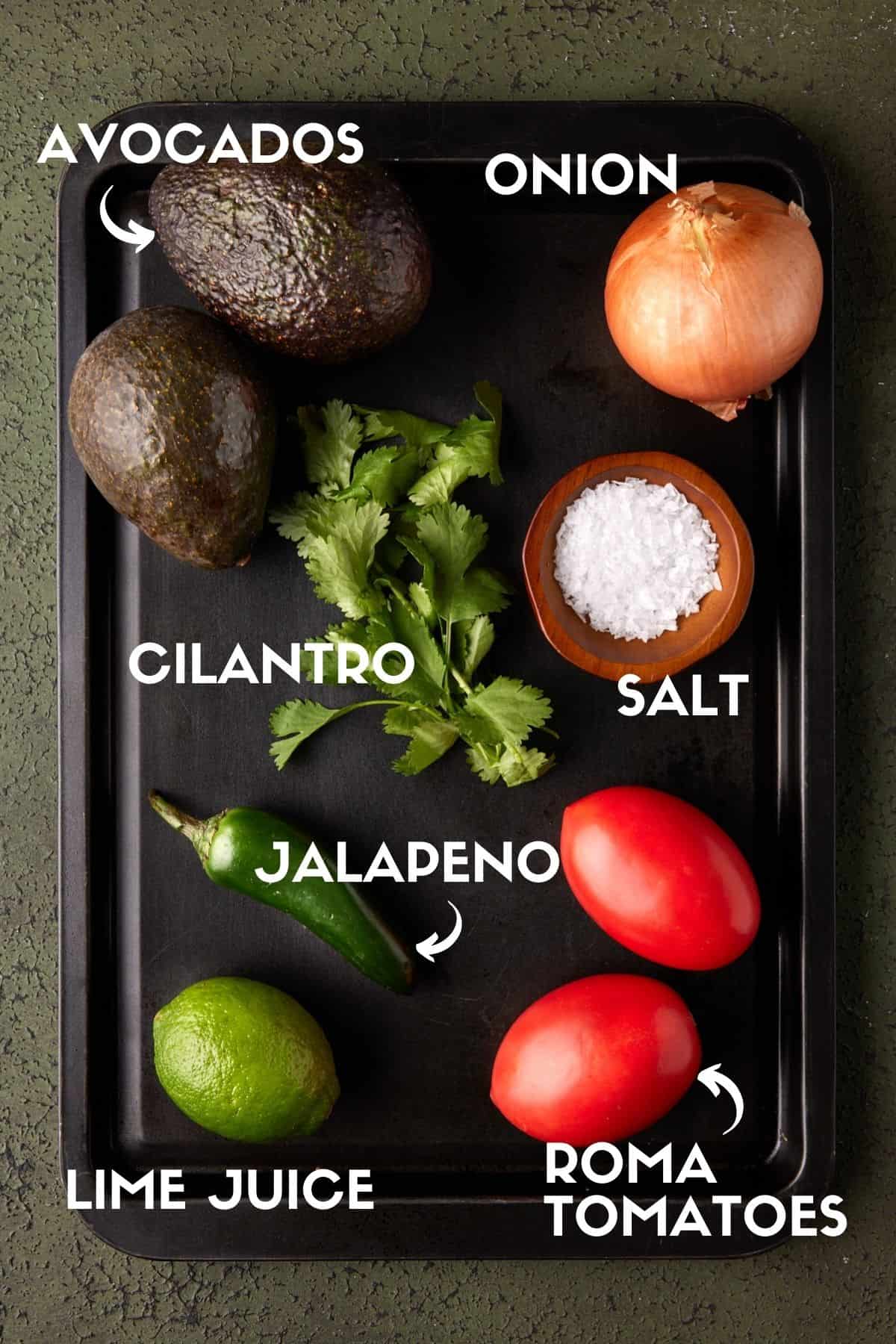 Guacamole ingredients on black sheet pan including avocados and tomatoes.