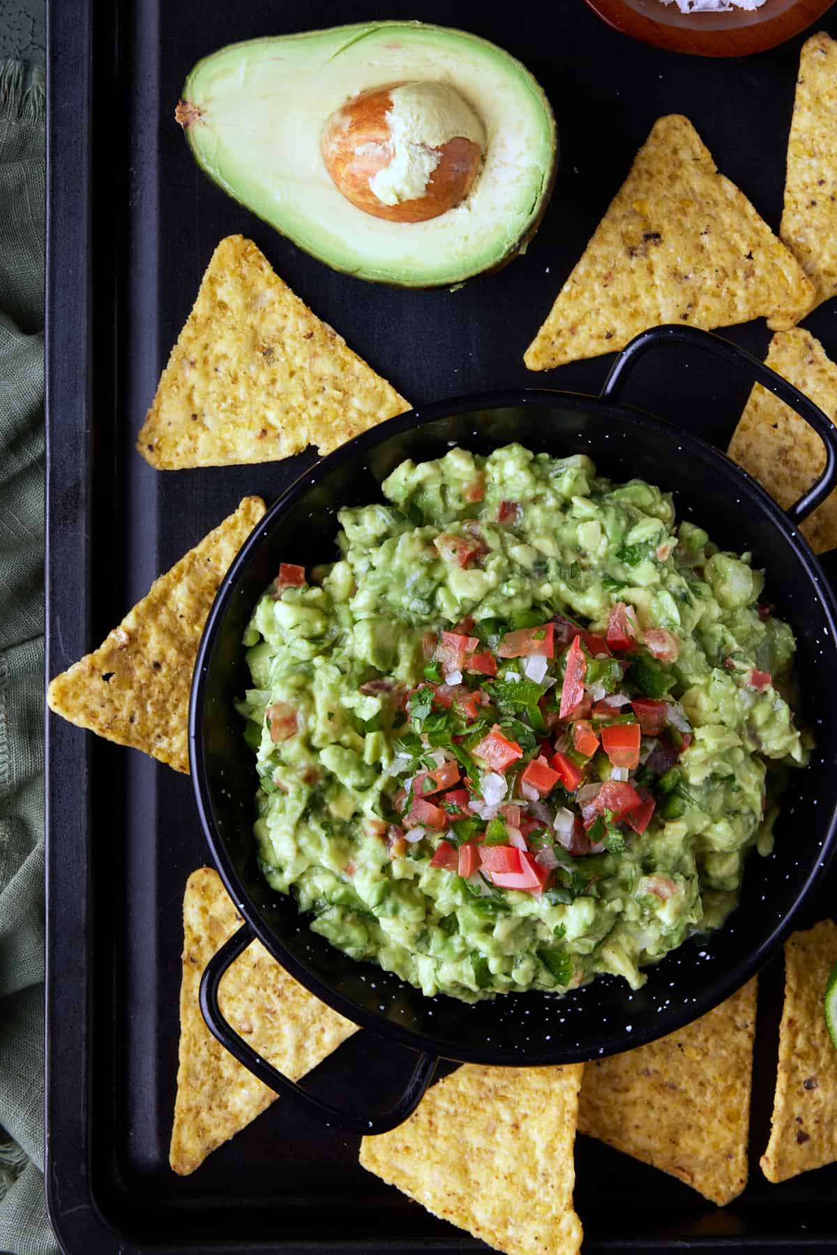 Black dish of chunky guacamole on black sheet pan with tortilla chips. 