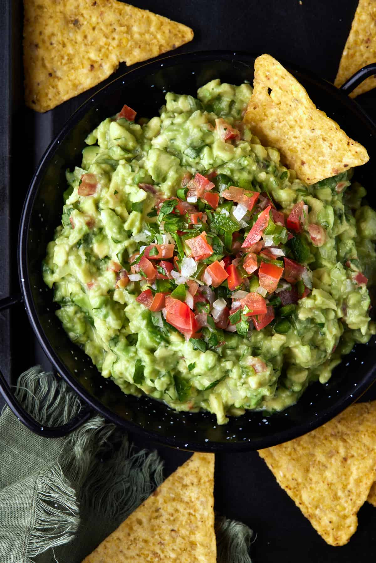 Black metal bowl filled with chunky guacamole and surrounded by tortilla chips. 