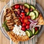 chicken souvlaki skewers in a bowl with pita, hummus and feta cheese.