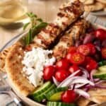 Chicken Souvlaki skewers with feta cheese, pita and hummus in a bowl with fresh vegetables.