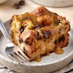Slice of chocolate croissant bread pudding with vanilla sauce on cream plate with fork.