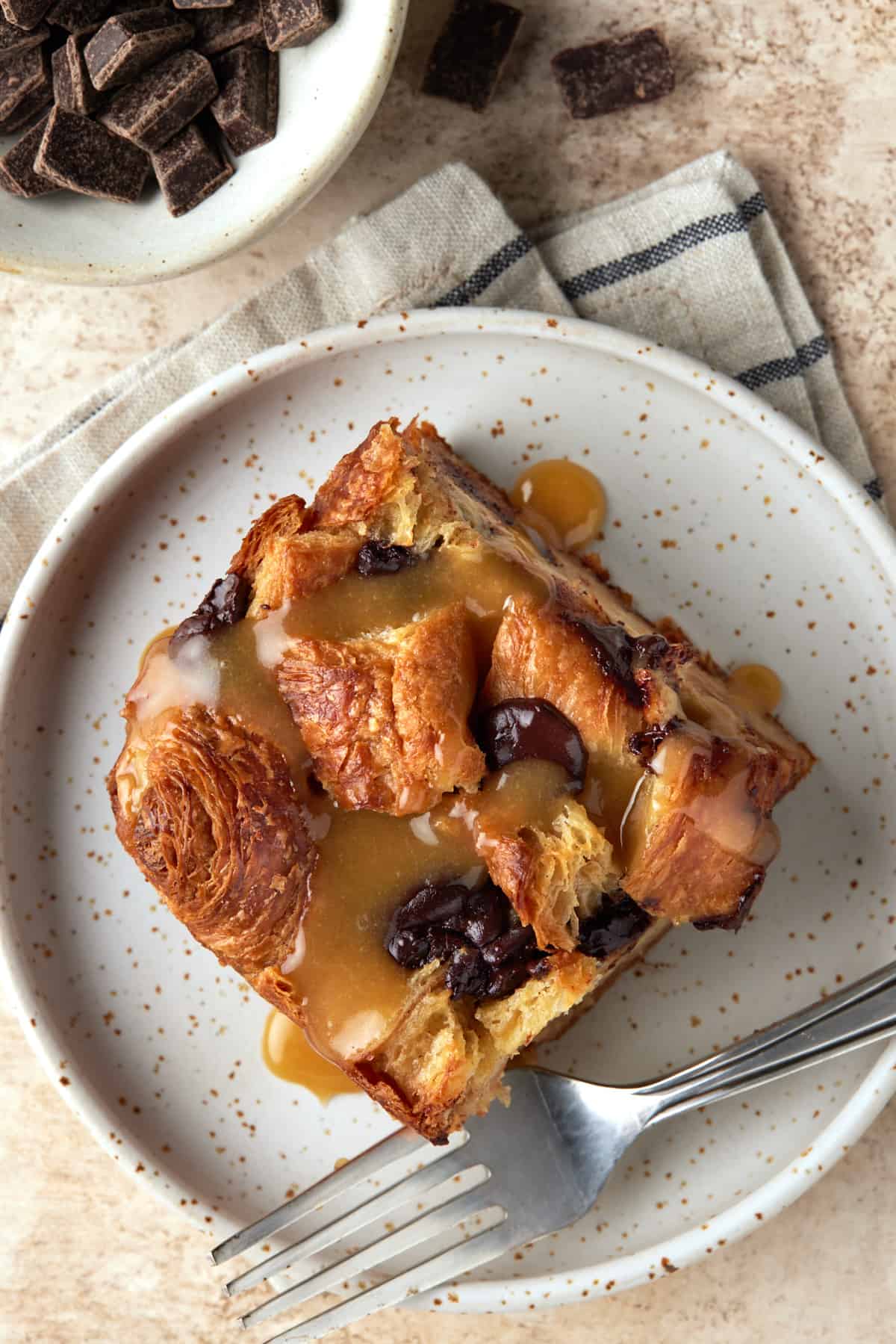 Square of baked Chocolate Croissant Bread Pudding with Vanilla Sauce on a cream plate with a fork.