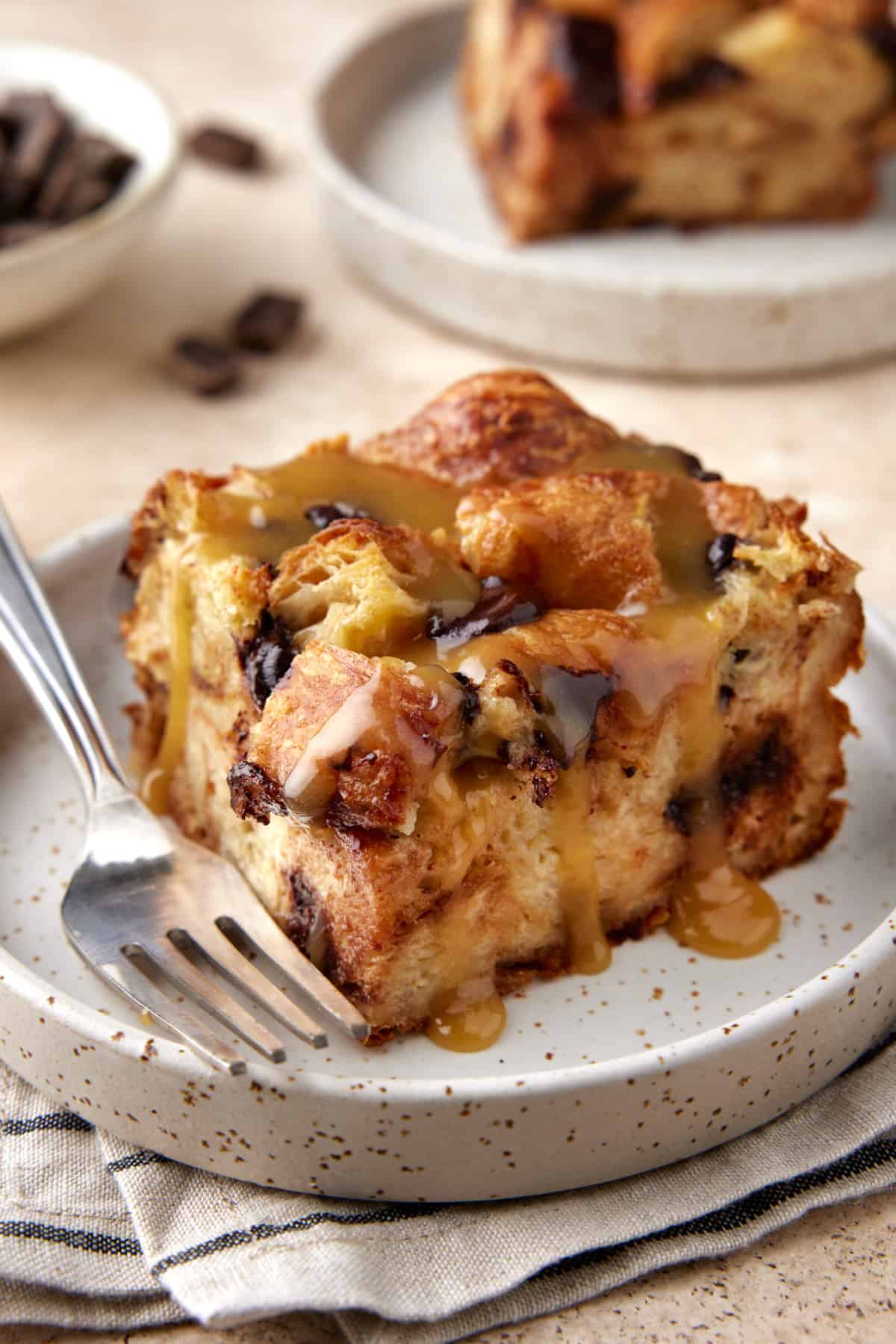 Slice of croissant bread pudding on cream plate with sauce and fork. 