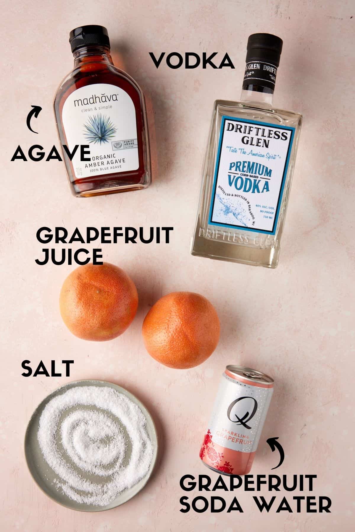 Bottles of vodka, agave, grapefruit soda, a grapefruit and salt on a pink board. 