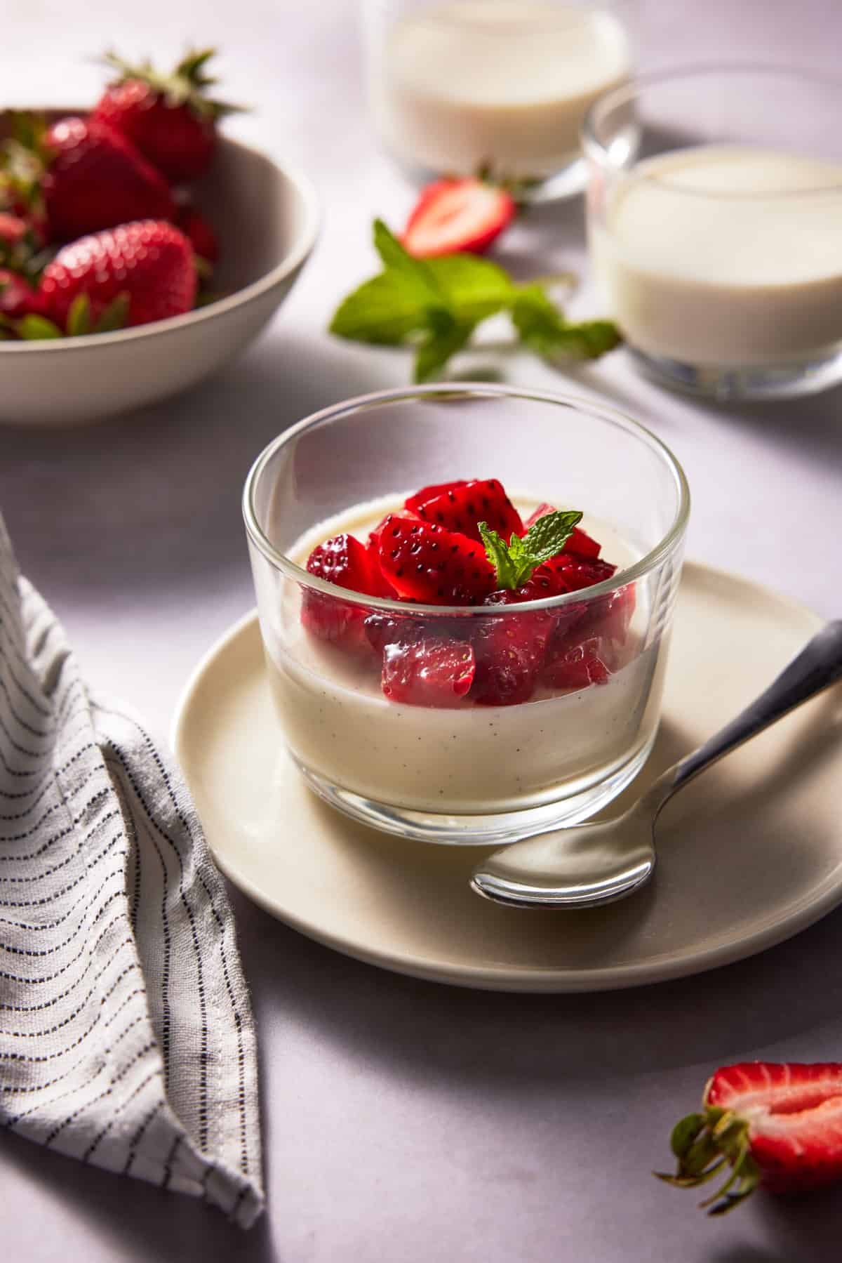 Glass ramekin with vanilla bean panna cotta and chopped strawberries on a cream plate. 