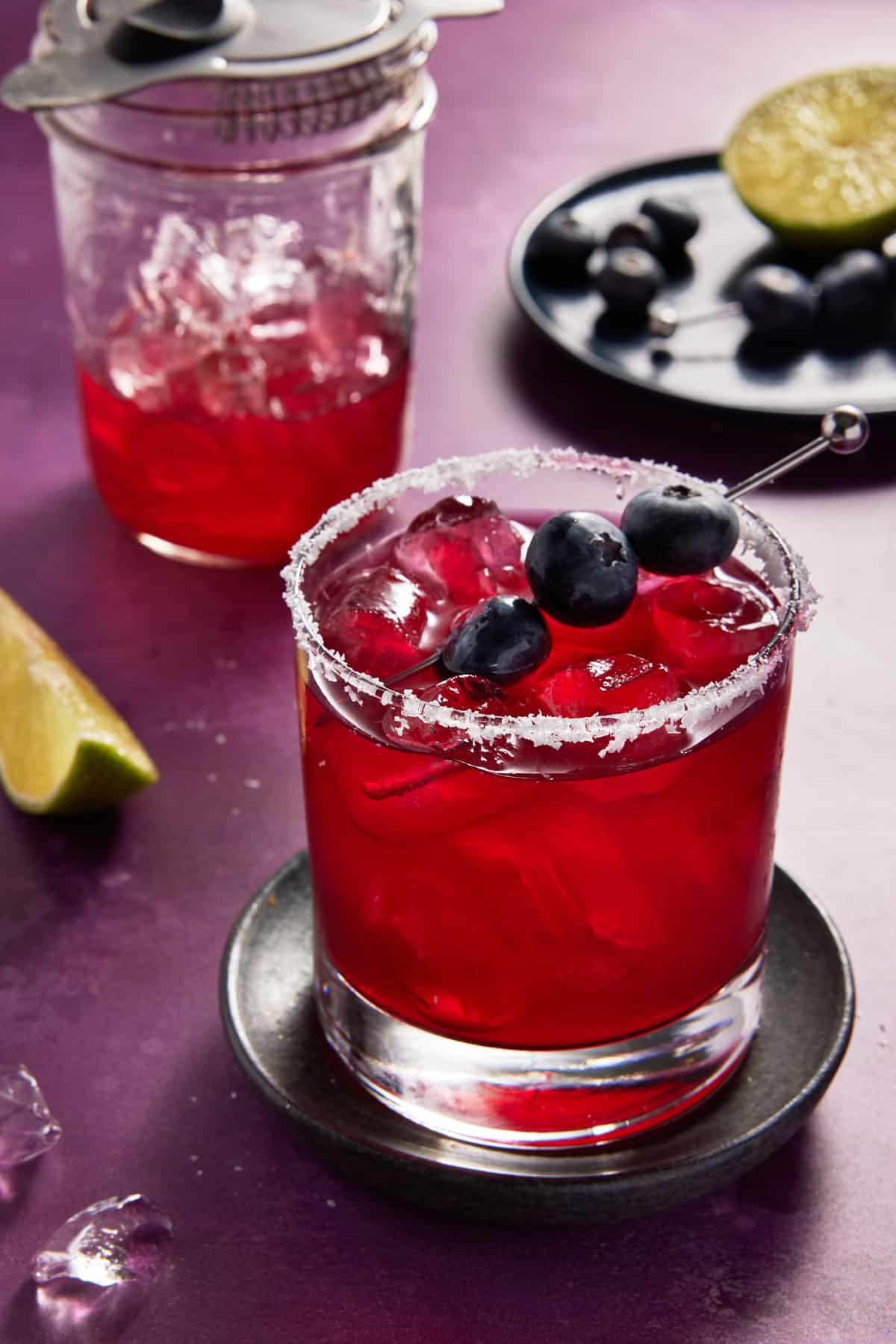 Low ball salt rimmed glass filled with blueberry margarita and a shaker filled with ice and margarita in the back.