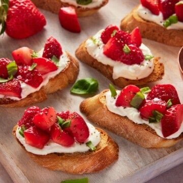 Bruschetta topped with strawberries, goat cheese and basil on a light wood cutting board.