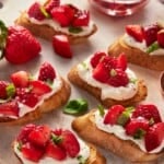 Slices of bruschetta topped with goat cheese and strawberries on a light wood cutting board.