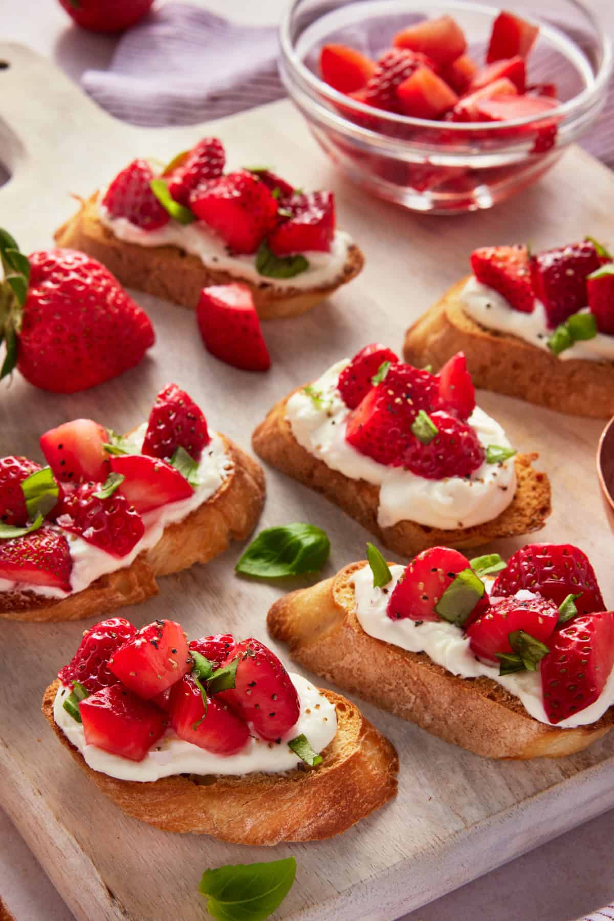 Sliced pieces of Goat cheese bruschetta topped with strawberries and basil on a light wood cutting board. 