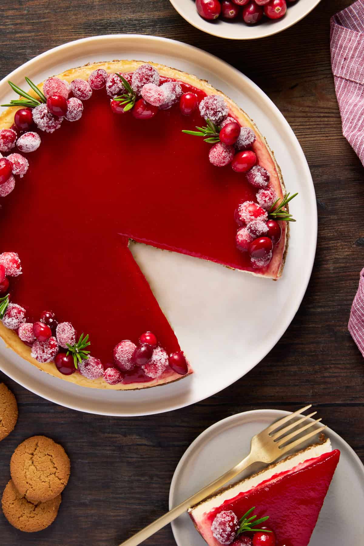 Cranberry cheesecake garnished with sugared cranberries on a white plate with a slice removed. 