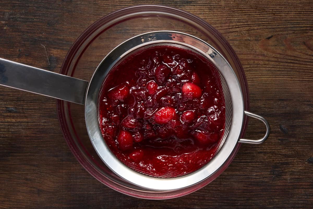 Cooked cranberries in a metal sieve over a glass bowl. 