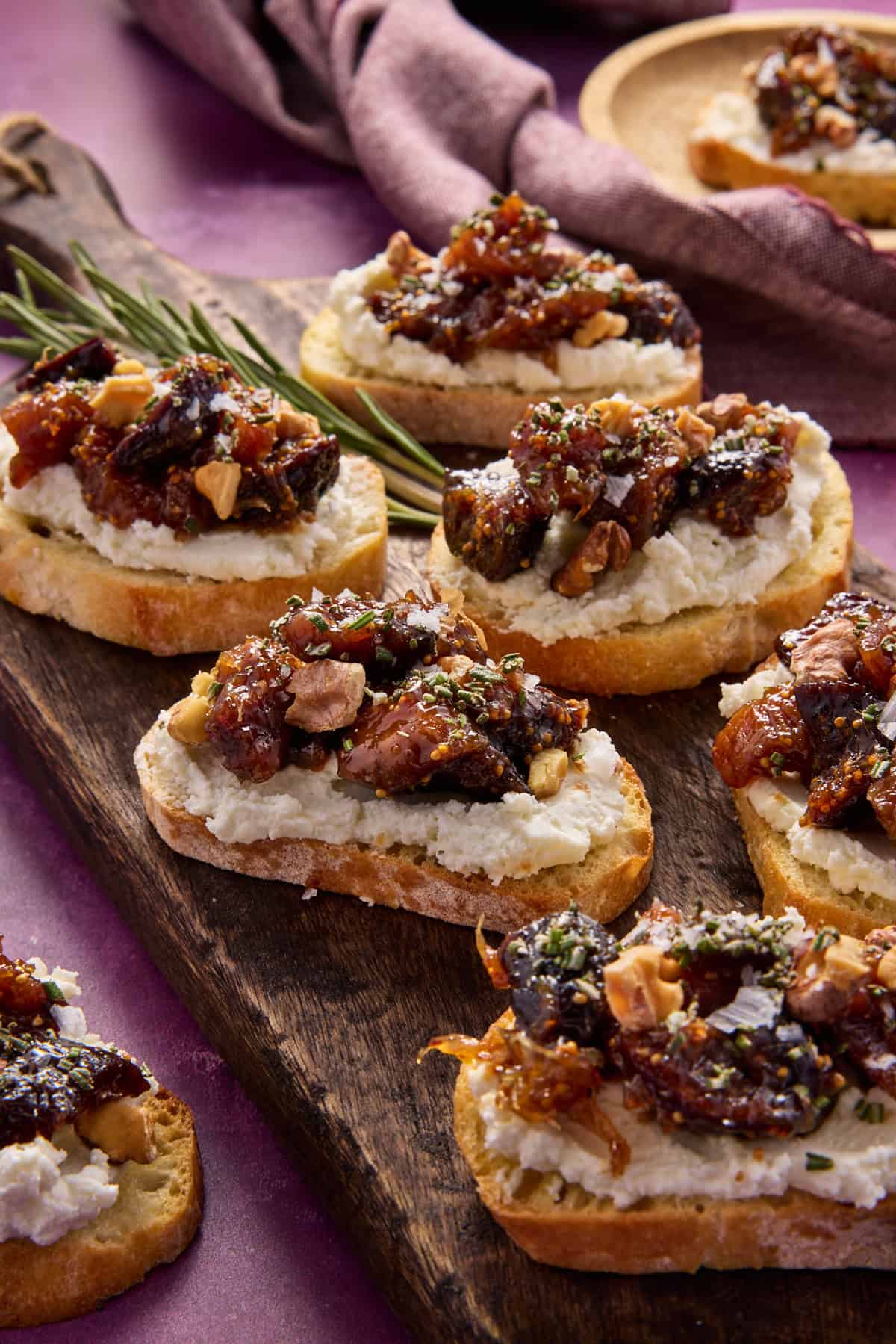 Fig and goat cheese crostini sprinkled with rosemary, walnuts and sea salt on a dark wood serving board. 