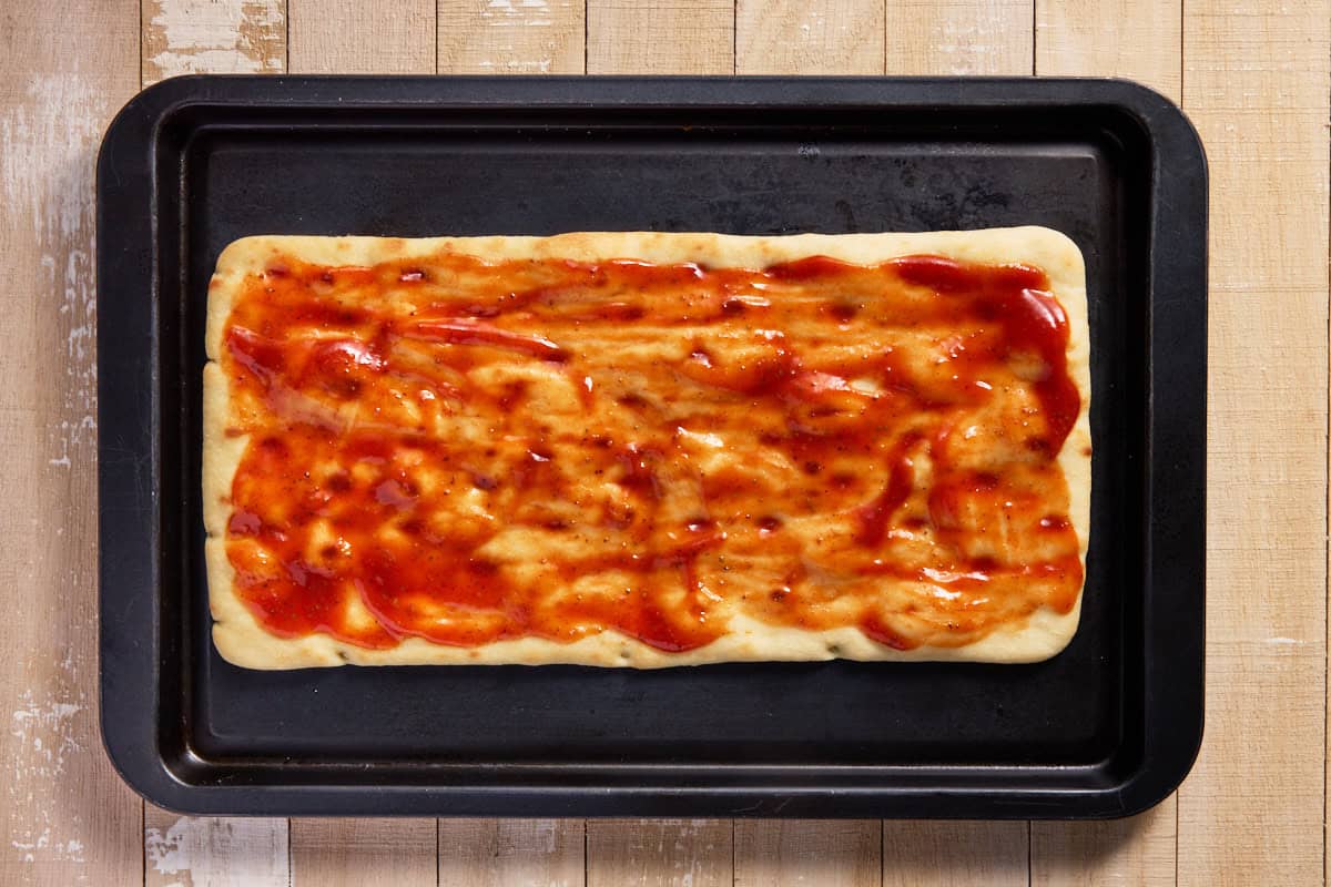 Flatbread crust with BBQ sauce on a black sheet pan. 