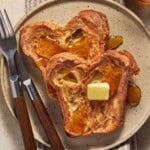 Two pieces of croissant bread french toast with butter and syrup on a beige plate.