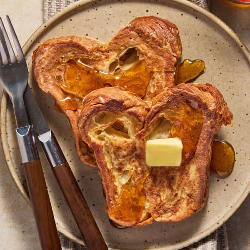 Two pieces of croissant bread french toast with butter and syrup on a beige plate.