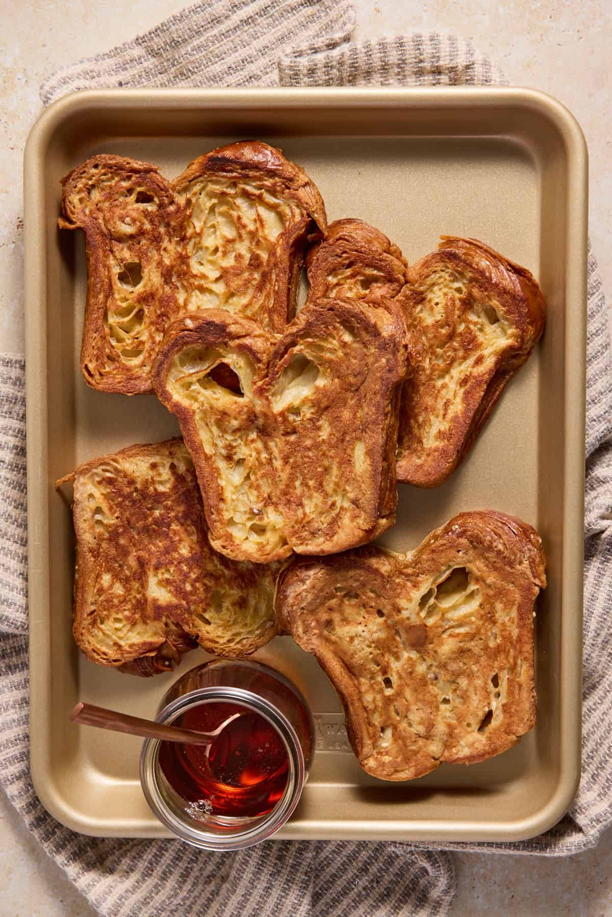 Pieces of croissant bread french toast on a sheet pan with a jar of maple syrup. 