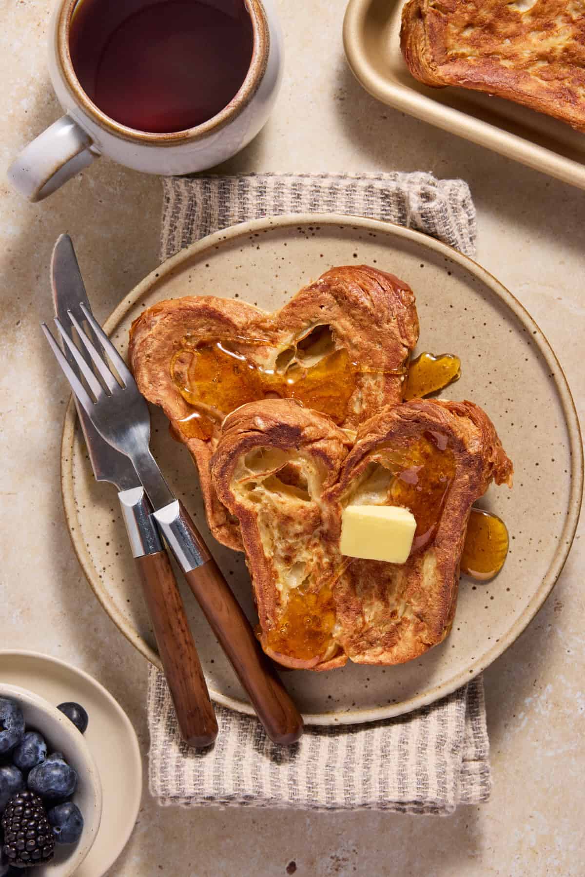 Two slices of croissant bread french toast on a beige plate with butter and syrup.