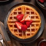 Round Belgian style yeast waffle topped with sliced strawberries and powdered sugar on a dark plate.
