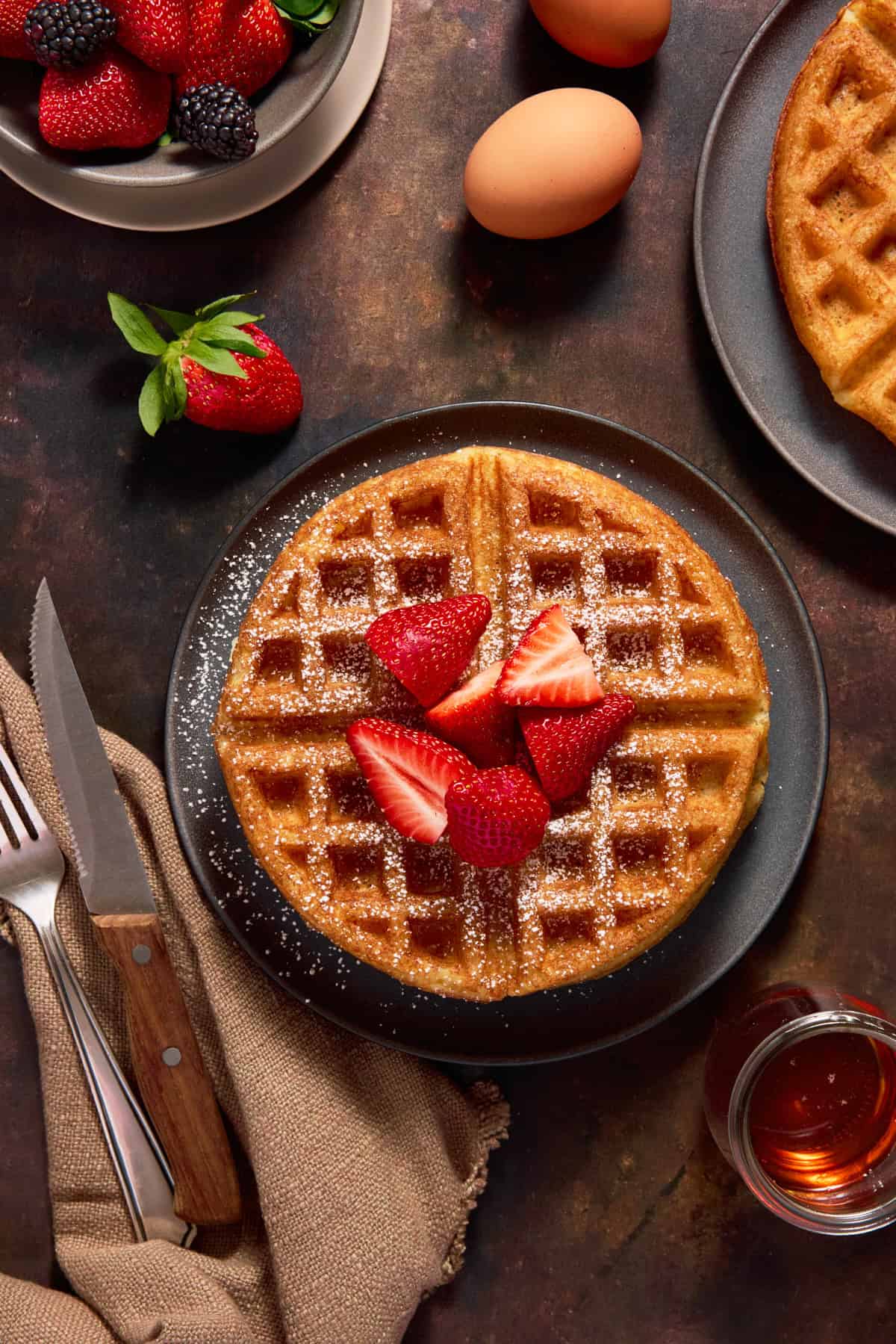 Overnight Yeast Waffle topped with strawberries and powdered sugar on a dark plate. 