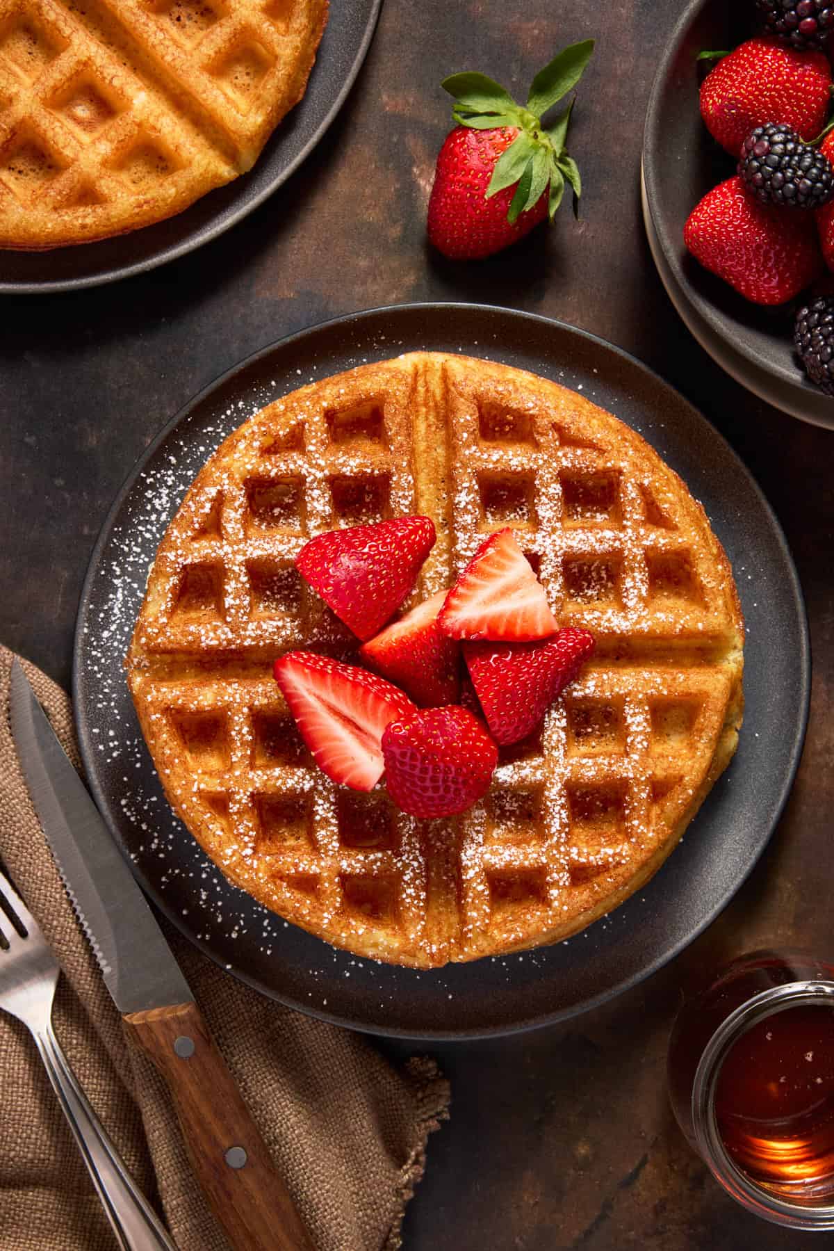 Round yeast waffle topped with sliced strawberries on a dark round plate.