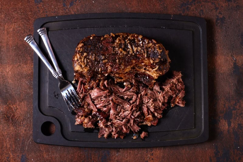 Chuck roast being shredded with two forks on cutting board.