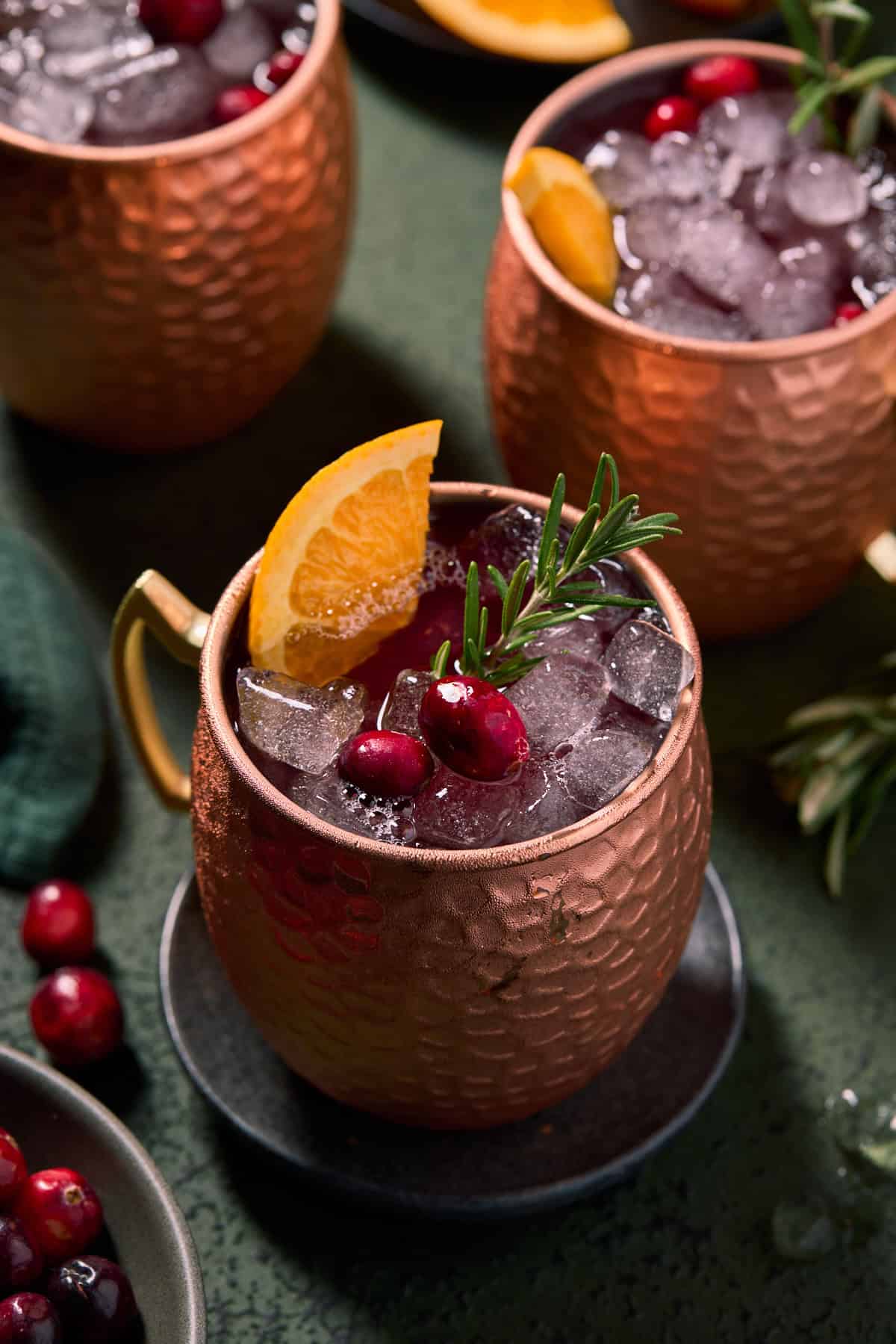 3 cranberry Moscow mules in copper mugs with fresh fruit for garnishes.