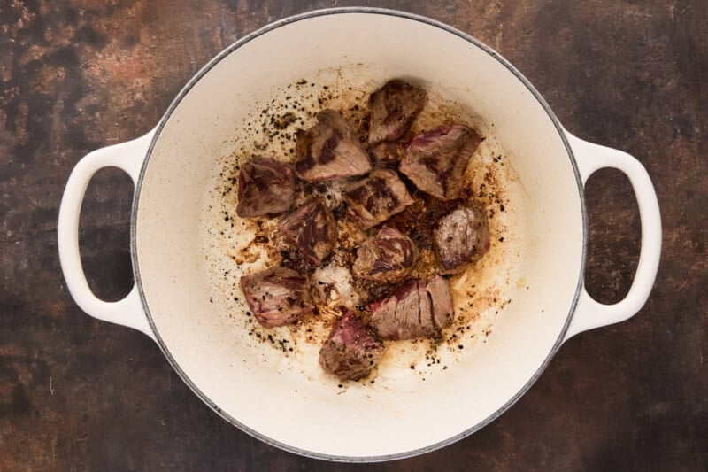 Pieces of beef seared in a white dutch oven.