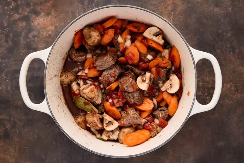 Seared beef in white dutch oven with carrots, mushrooms, tomatoes, onions, garlic, fresh herbs and red wine.
