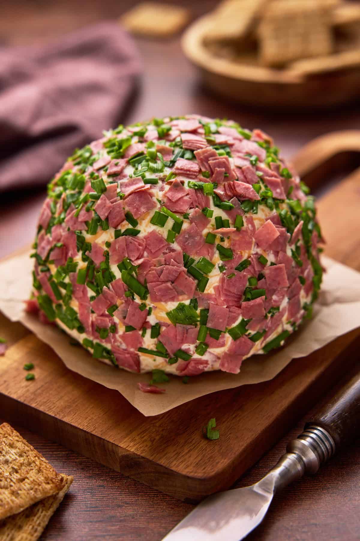 Cheese ball garnished with dried beef and chopped chives on a brown board with a spreader.