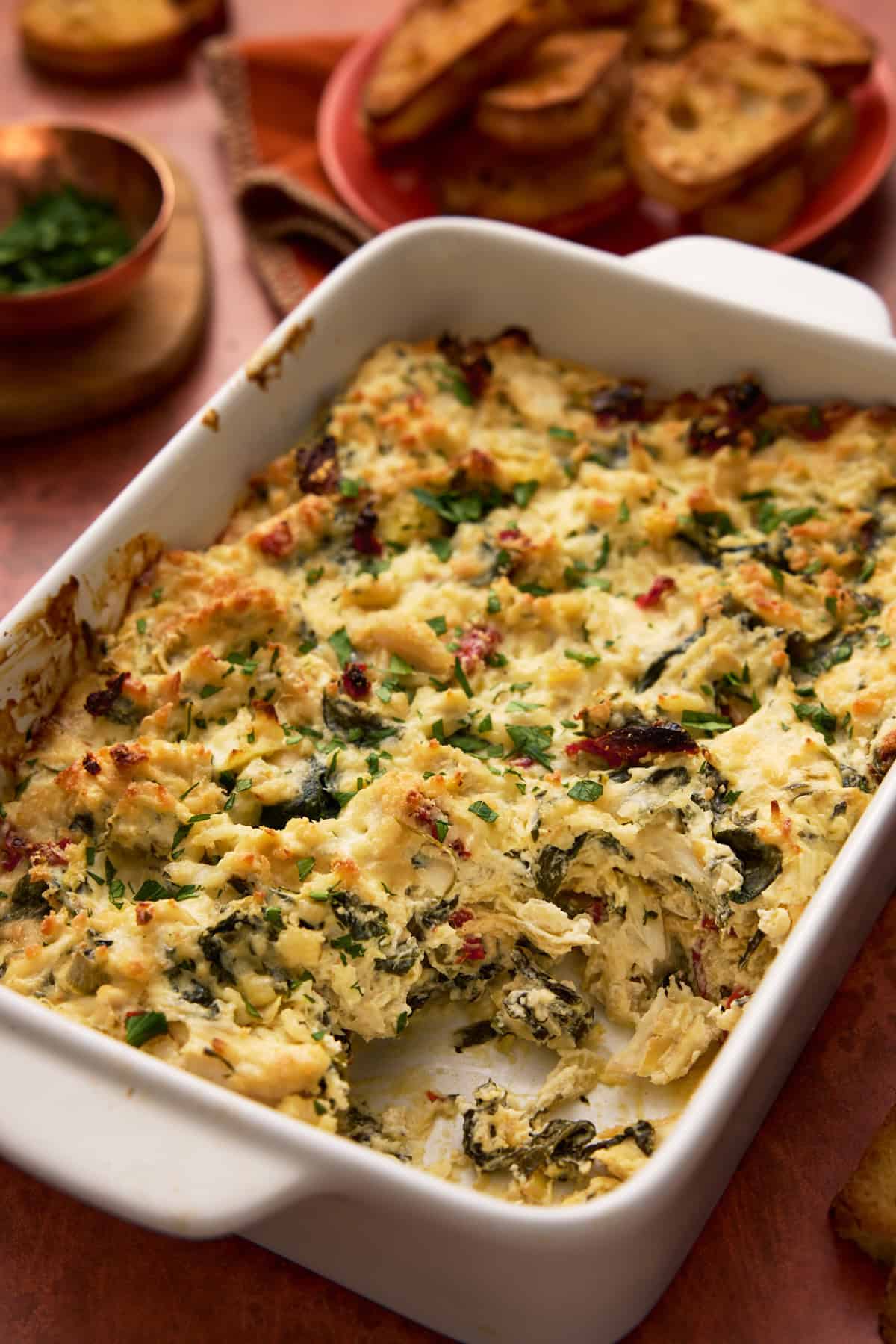 Crab artichoke dip with sun-dried tomatoes and spinach in white baking dish.