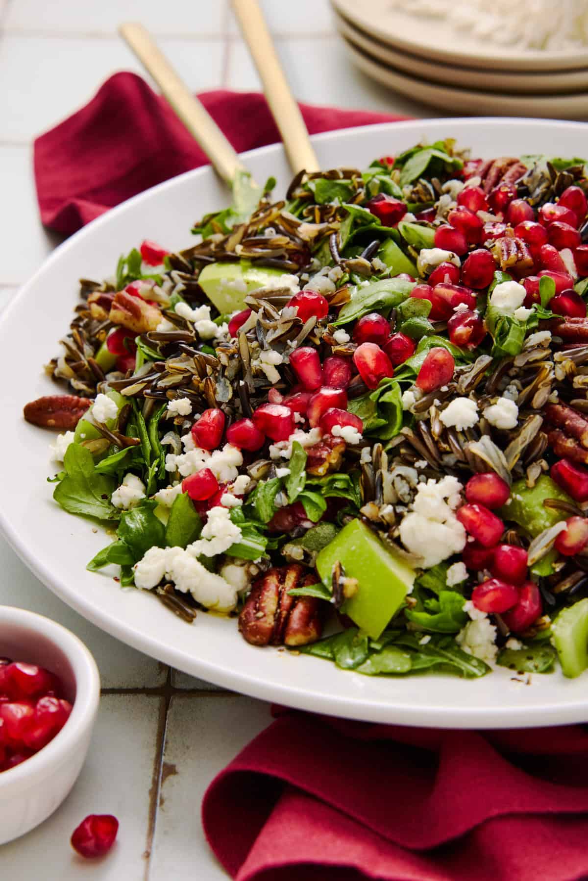 Side angle of a holiday wild rice salad with pomegranate, arugula and goat cheese on a white plate with gold serving spoons.