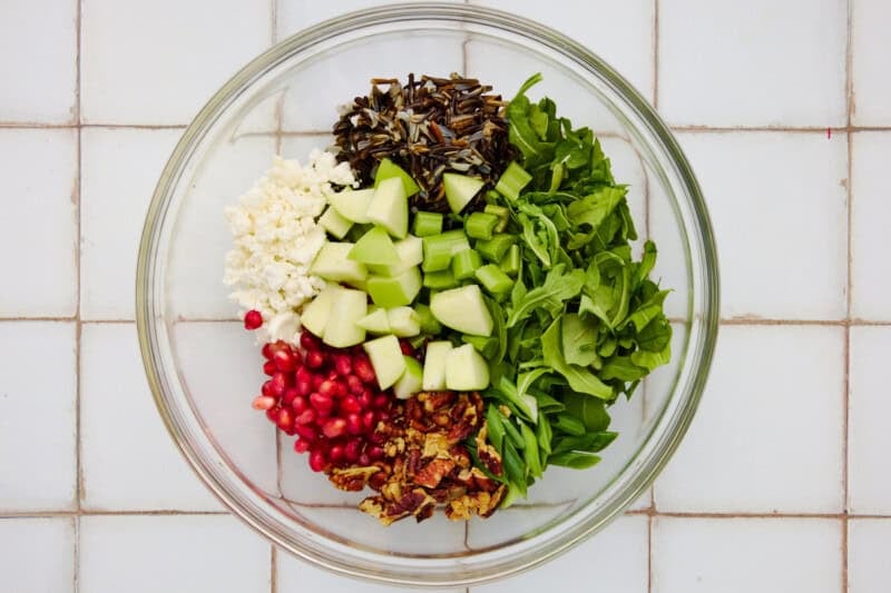 Holiday salad ingredients in a glass bowl including arugula, apples, celery, pomegranate, pecans, wild rice and goat cheese.