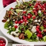 Holiday Salad with wild rice, pecans, goat cheese, apples and arugula on a white plate with a red napkin and gold serving spoons.