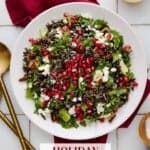 Holiday Salad with wild rice, pecans, goat cheese, apples and arugula on a white plate with a red napkin and a glass bowl of vinaigrette.