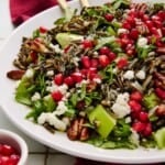 Holiday Salad with wild rice, pecans, goat cheese, apples and arugula on a white plate with a red napkin and gold serving spoons.