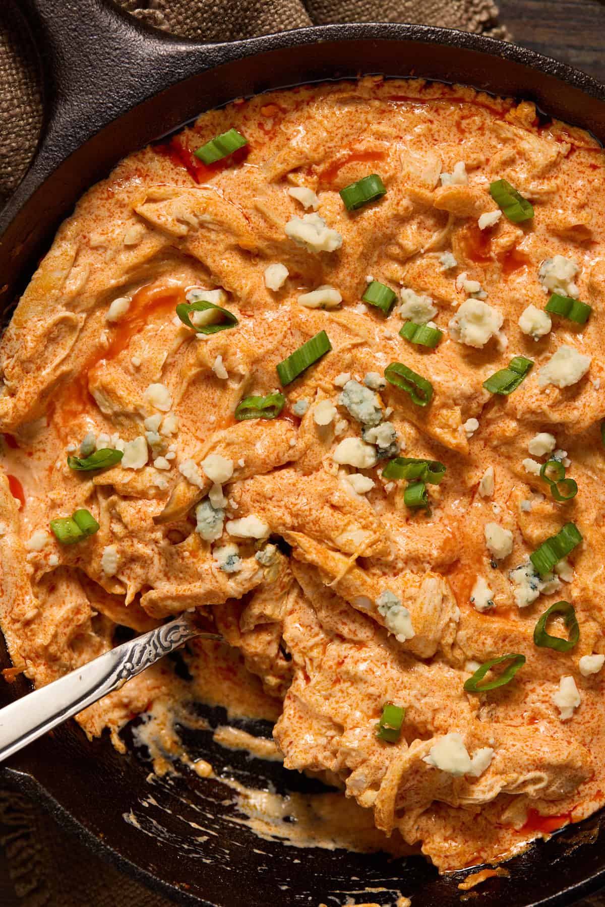 Cast iron pan of buffalo chicken dip with a spoon in it and garnished with blue cheese and scallions.