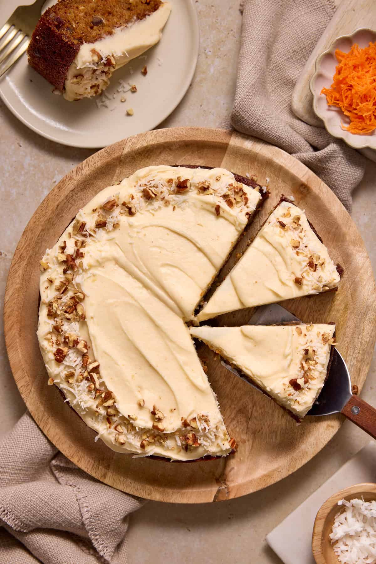 A single layer carrot cake topped with chopped pecans and shredded coconut on a wooden charger. Three slices have been cut and one put on a plate.