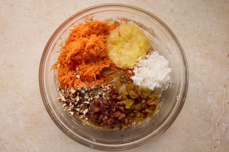 Ingredients for single layer carrot cake in a glass bowl, including grated carrots, pineapple, pecans and coconut.
