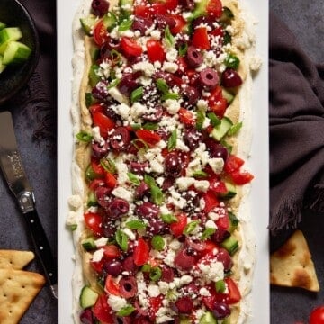 Layered Greek Dip on a white platter surrounded by pita chips.