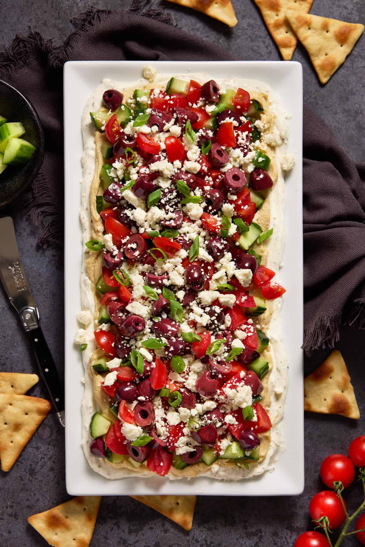 Layered Greek Dip on a white platter surrounded by pita chips, tomatoes and cucumbers.