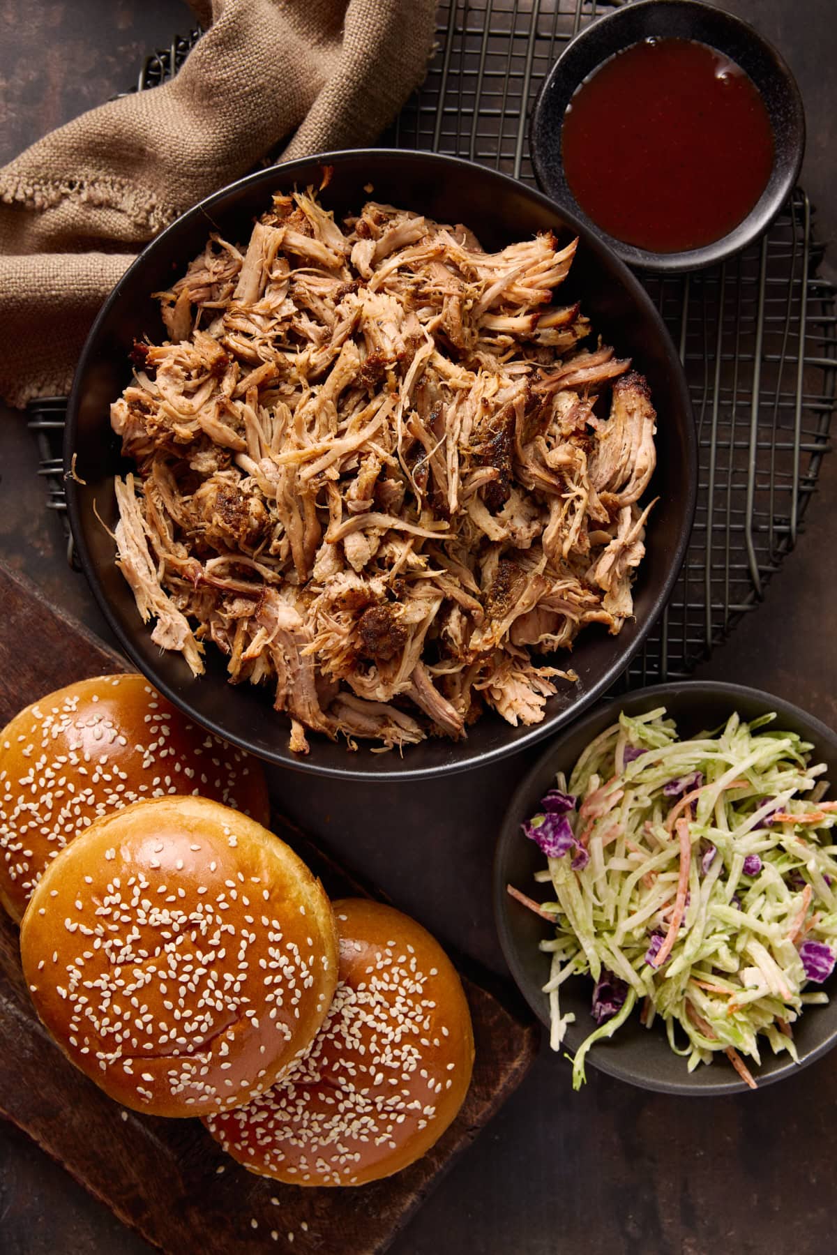 Bowl of pulled pork, pile of buns, bowl of bbq sauce and a bowl of broccoli slaw.