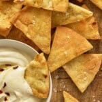 Air fryer pita chips on a wooden board along with a chip dunked in a bowl of hummus.